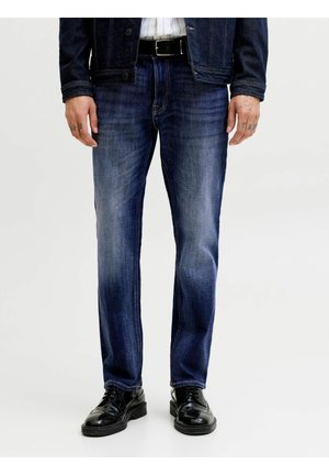 Mężczyzna w ciemnoniebieskich, wyblakłych dżinsach, czarnym pasku, czarnych butach sznurowanych oraz ciemnej kurtce stoi na tle jednolitego jasnego tła.