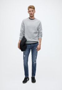 Šedá mikina s černým lemem, modré potrhané džíny a černé tenisky. Drží černou tašku, stojí před jednobarevným pozadím.