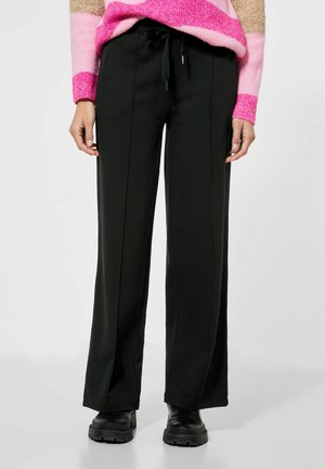 Pantaloni neri a gamba larga con una texture liscia, dotati di dettagli sulle cuciture laterali e una vita elastica con cordino. Abbinati a un pullover lavorato a maglia colorato.