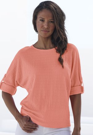 Koraalkleurige blouse met een grove textuur, een brede halslijn, driekwart mouwen opgerold en een relaxte pasvorm. Witte broek gedragen, handen op de heupen.