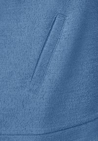 Niebieska teksturowana tkanina z subtelnym wzorem, z małym, skośnym detalem kieszeni. Materiał wydaje się miękki i tkany.