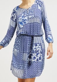 Robe bleue à motifs avec manches longues et taille cintrée. Présente un mélange de motifs cachemire et géométriques, agrémentée d'un cordon de serrage et de franges.