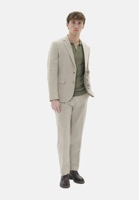 Beige Leinenanzug mit einer taillierten Jacke und passender Hose, kombiniert mit einem grünen strukturierten Polohemd. Braune Schnürschuhe vervollständigen den Look.