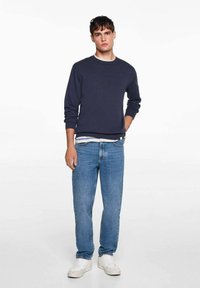 Marinasti pulover s posadnimi ovratnikom, bombažno mešanico, oblečen čez belo in mornarsko modro črtasto srajco. Svetlo modri džinsi in beli športni čevlji zaključujejo obleko.