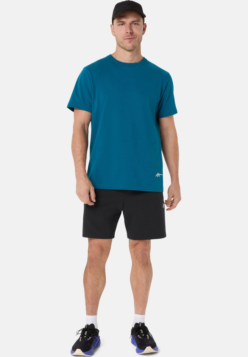 T-shirt en coton bleu sarcelle avec col rond, porté avec un short noir. Baskets foncées avec des accents bleus. Design uni, sans motifs visibles.