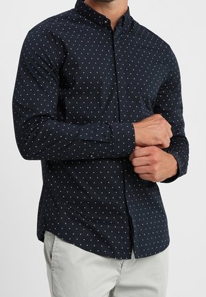 Man wearing a fitted navy blue button-up shirt with small white triangle patterns and light gray pants, adjusting his cuff.