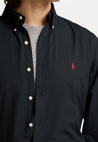 Svart skjorta med knappar och spetsig krage, vita knappar och en liten röd polo-logga på bröstet, överlagd på en grå crewneck-tröja.