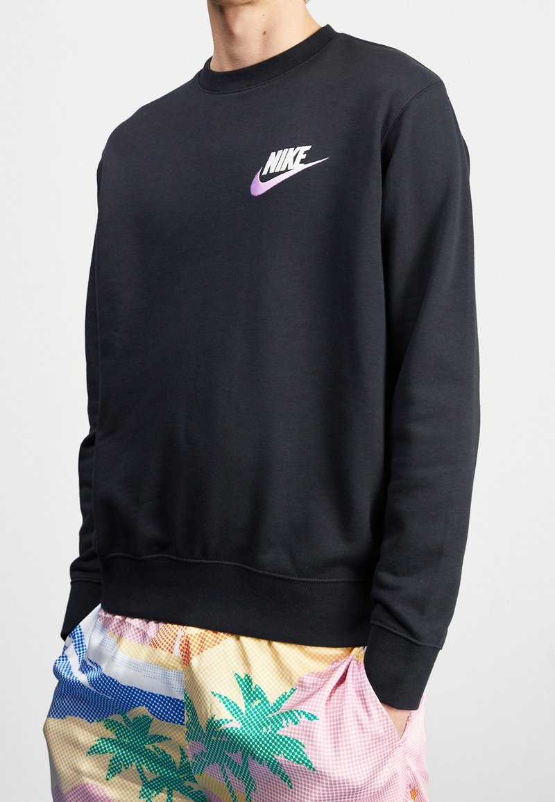 Personne portant un sweat-shirt Nike noir avec un logo swoosh violet et un short coloré à motifs de palmiers.