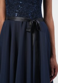 Navy dress featuring a lace bodice with sequins, a flowing skirt, and a satin black ribbon belt tied at the waist. Smooth texture overall.