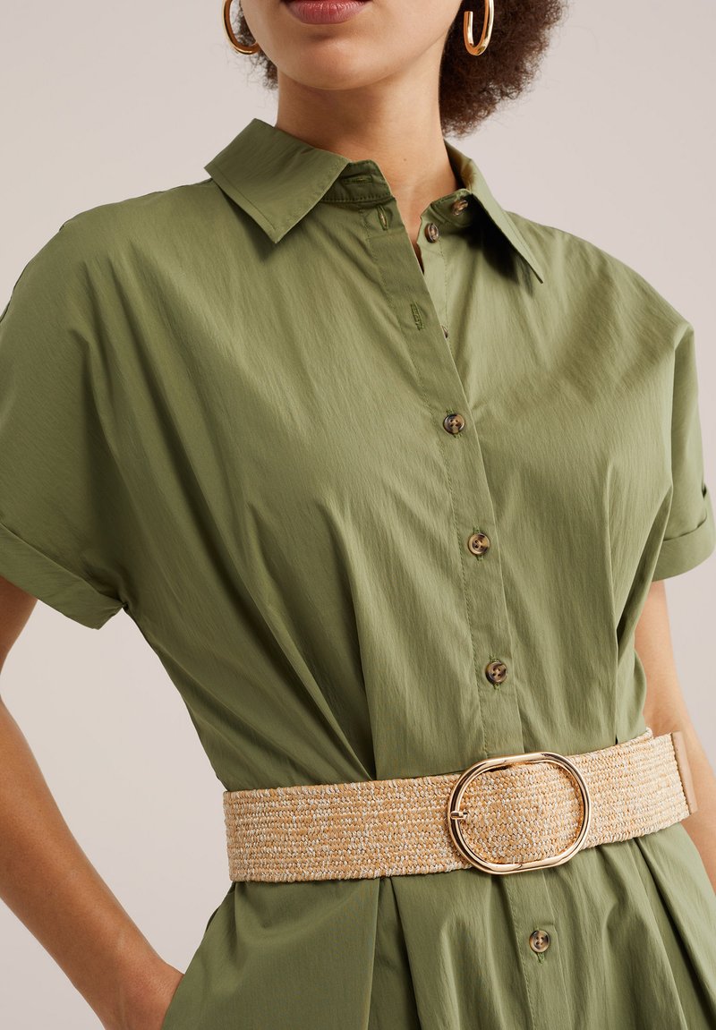 Femme portant une robe boutonnée vert olive aux manches retroussées, de grandes boucles d'oreilles créoles dorées et une large ceinture tissée beige avec une boucle ovale.