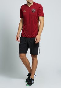 Röd sporttröja med v-halsringning, prydd med en gyllene örn-emblem och gyllene detaljer. Matchas med svarta shorts och svarta löparskor.