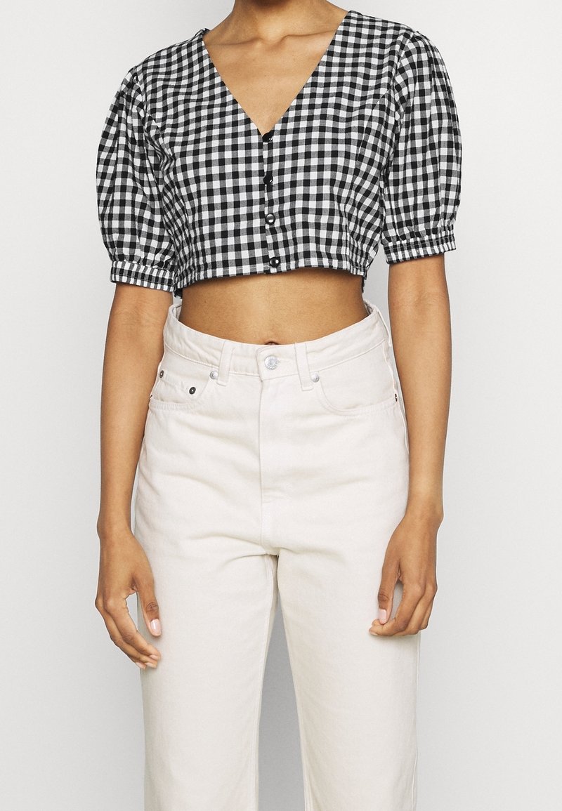 Blouse courte à carreaux noir et blanc avec un décolleté en V plongeant et des manches bouffantes, ornée de boutons noirs sur le devant, associée à un pantalon clair.
