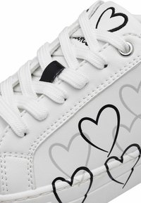 Baskets en cuir blanc avec des graphiques de cœurs noirs, des lacets blancs, une surface texturée et une bordure cousue le long de la semelle.