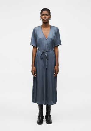 Mujer de pie de frente, vistiendo un vestido midi azul con mangas cortas, escote en V, cinturón atado y botas negras hasta la mitad de la pantorrilla sobre un fondo liso.