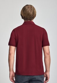 Camisa polo burdeos de tela texturizada, con mangas cortas y un cuello clásico. La vista trasera muestra un dobladillo limpio y costuras suaves.