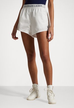 Vita sportshorts i vitt med dragsko i midjan och en marinblå logotyp, i kombination med vita höga sneakers. Slät textur, avslappnad design.