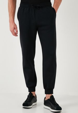 Schwarze Sweatpants mit elastischem Bund, Seitentaschen und Bündchen an den Knöcheln. Aus weichem Stoff gefertigt, kombiniert mit schwarzen Sportschuhen.