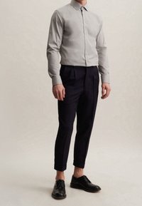 Ljusgrå långärmad skjorta med knappfront, kombinerad med mörkblå plisserade byxor och svarta finskor, stående mot en neutral bakgrund.