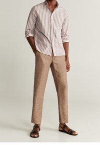 Camisa de botones a rayas en beige claro y franjas más oscuras, combinada con pantalones de lino marrón y sandalias de cuero marrón.