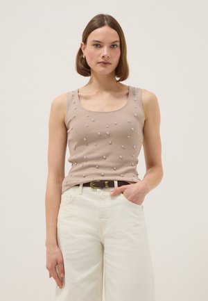 Mujer con cabello corto y castaño que lleva una blusa sin mangas beige con pequeñas perlas, pantalones blancos de talle alto y un cinturón oscuro, de pie frente a un fondo liso.