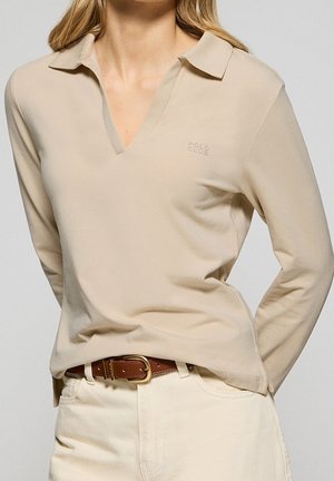 Vrouw die een beige poloshirt met lange mouwen draagt, met een klein "Polo Club"-logo, ingestopt in crèmekleurige broek met een bruine leren riem.