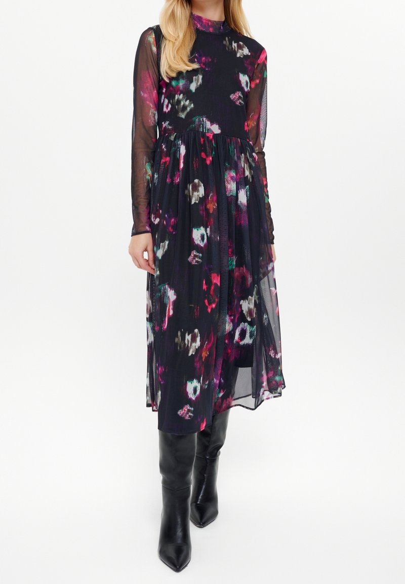Femme portant une robe midi noire transparente à manches longues avec des imprimés floraux abstraits colorés et des bottes noires montantes à bout pointu.