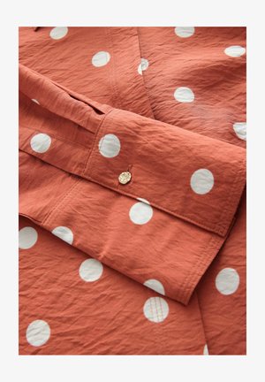 Hnědo-oranžová látková košile s bílými puntíky, se skládaným manžetem a detailem zlatého knoflíku, ukazující texturovaný povrch.