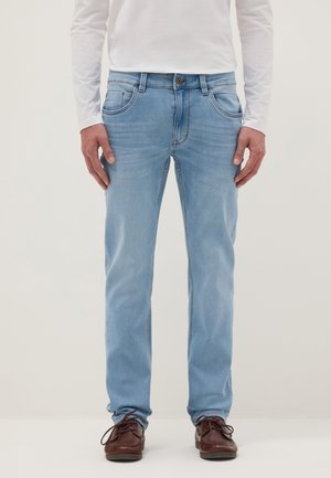 Hombre vistiendo jeans azul claro, camisa blanca de manga larga y zapatos de cuero marrón, de pie contra un fondo liso.