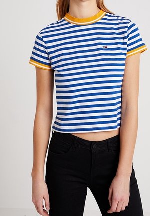 Persoon in een blauw-wit gestreepte crop top met gele details en zwarte broek, staande tegen een egale achtergrond.