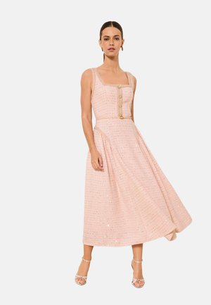 Frau trägt ein ärmelloses, blassrosa strukturiertes Midikleid mit goldenen Knöpfen und schmalem Gürtel, kombiniert mit Riemchen-Hochsandalen.