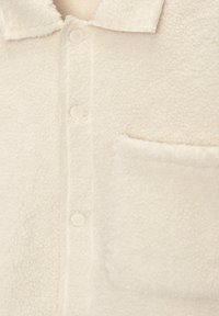 Veste en polaire sherpa couleur crème à l'avant avec boutons-pression, col pointu et poche poitrine sur le côté droit.