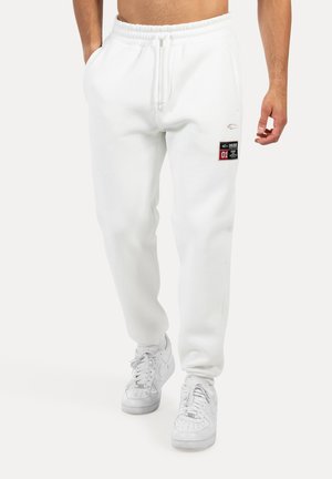 Joggers blancs en polaire avec une taille élastique, des poches latérales et un patch en label noir sur la cuisse, associés à des chaussures de sport blanches.
