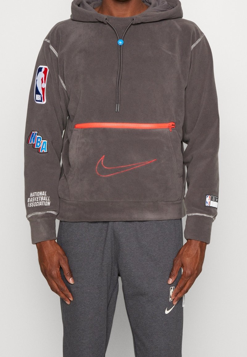 Felpa grigia in pile con cappuccio, dotata di tasca frontale con zip arancione, logo Nike e branding NBA sulle maniche; texture morbida.