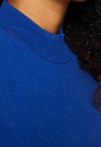 Kungligt blå mocknecktröja med en mjuk textur, med ribbade detaljer vid kragen och axeln. Närbild av tyget.