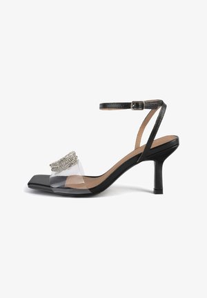 Sandal nera con tacco alto e cinturino trasparente, decorata con un accento testurizzato argento. Punta quadrata, sottile cinturino alla caviglia e tacco basso.
