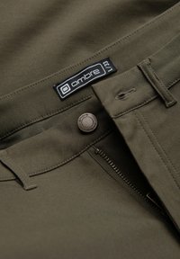 Gros plan d’un pantalon vert olive avec fermeture éclair ouverte, bouton et passants de ceinture ; étiquette de marque noire et blanche « ombre » visible sur la ceinture.