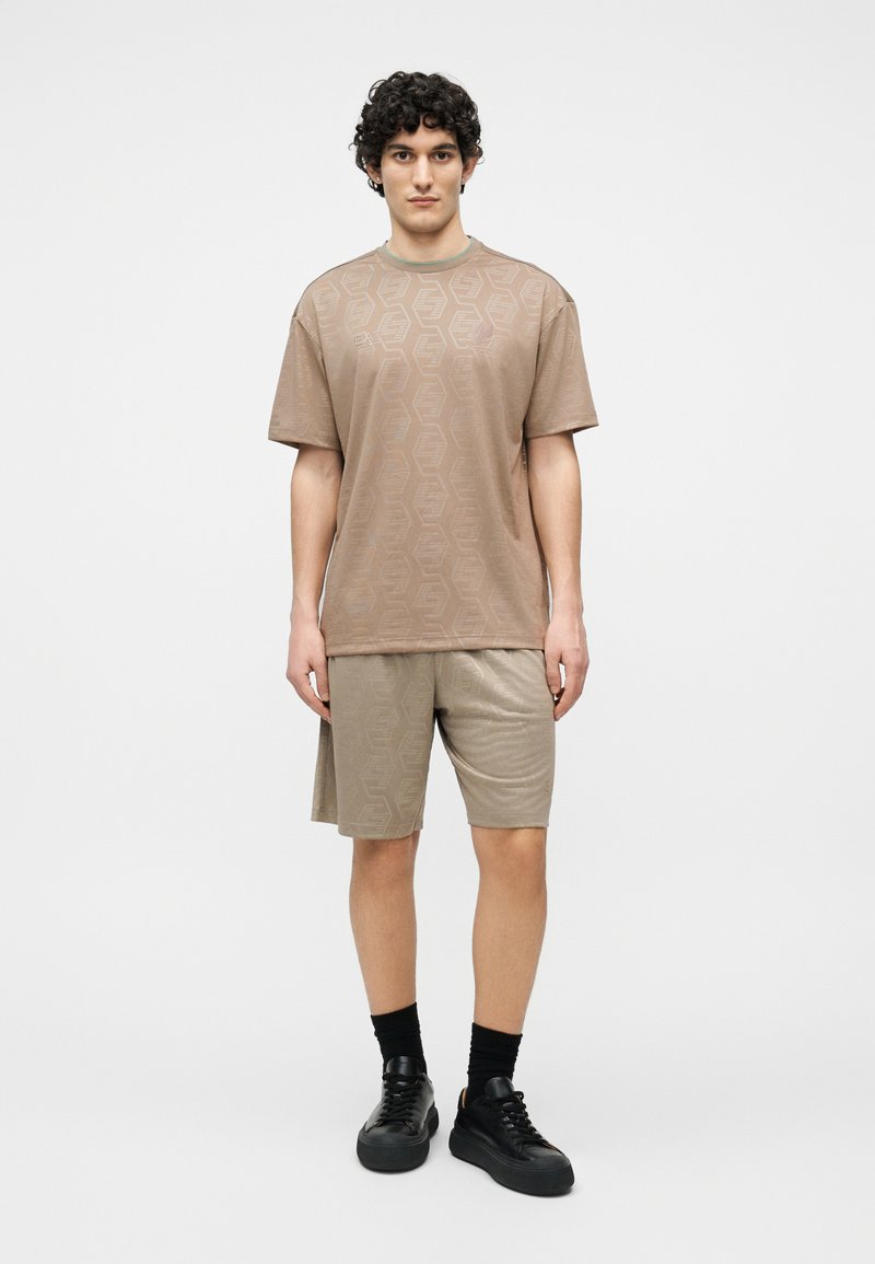 Νεαρός άνδρας με σγουρά μαλλιά φοράει ταιριαστό μπεζ ενισχυμένο t-shirt και σορτς με εξάγωνα μοτίβα, μαύρες κάλτσες και μαύρα αθλητικά παπούτσια, όρθιος.