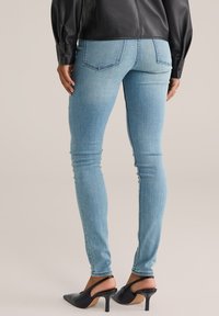 Žena v svetlomodrých skinny džínsoch, čiernej koženej blúzke a čiernych sandáloch na podpätku stojí na neutrálnej pozadí.