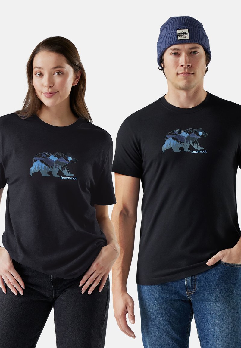 Schwarze Baumwoll-T-Shirts mit einem Grafikdruck eines Bären und einem blauen Bergmuster. Kombiniert mit dunklen Jeans und einer blauen Mütze.
