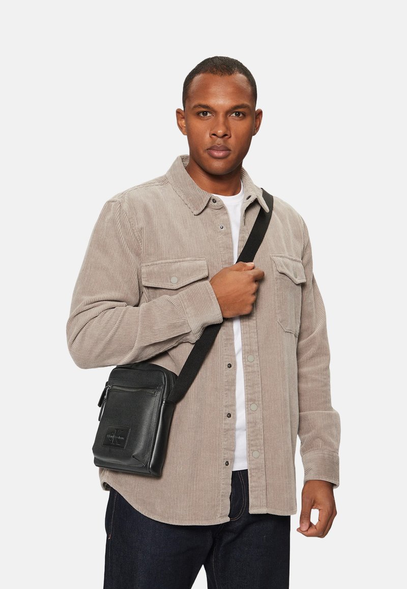 Bolso de hombro de cuero negro con cierre de cremallera, correa ajustable y un bolsillo frontal, modelado por una persona con una camisa de pana beige.