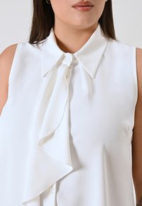 Blusa bianca senza maniche con colletto a punta, caratterizzata da un dettaglio a fiocco frontale e un tessuto liscio e lucido. Chiusura con bottoni visibile.