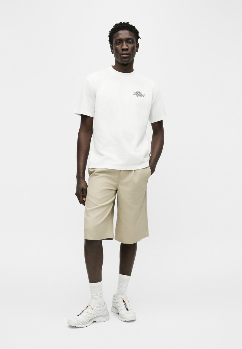 Homme debout, portant un t-shirt blanc à manches courtes avec un petit logo, un short beige arrivant au genou, des chaussettes blanches et des baskets blanches, main dans la poche.