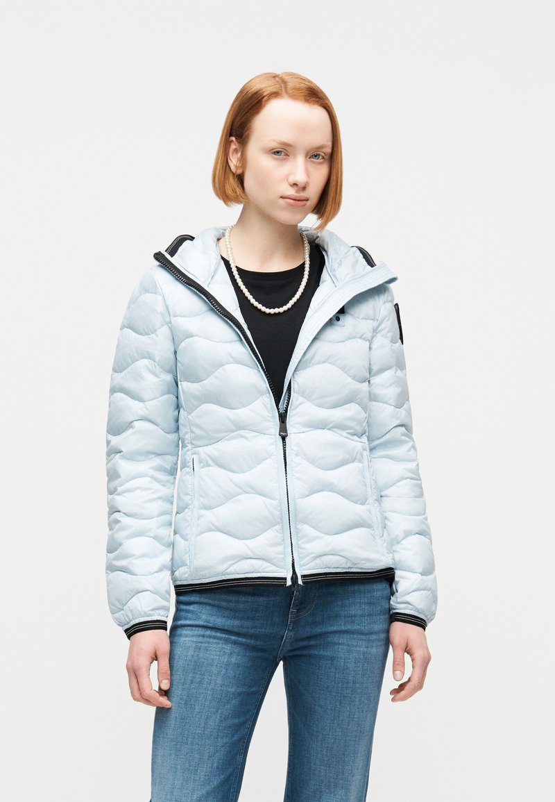 Νεαρή γυναίκα που φοράει ανοιχτό μπλε μπουφάν με καπιτονέ πάνω από μαύρο τοπ με πέρλες και μπλε τζιν, στέκεται μπροστά σε λευκό φόντο.