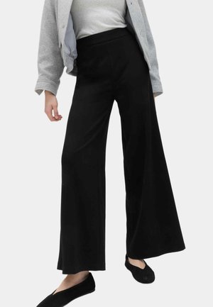 Persona con pantalones anchos negros, chaqueta gris, camiseta blanca y zapatos planos negros, de pie contra un fondo claro y liso.