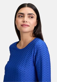 Blaues, strukturiertes Oberteil mit einem Wabenmuster, rundem Ausschnitt und langen Ärmeln. Glattes Material mit leichtem Glanz und tailliertem Schnitt.