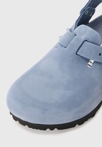 Zoccolo in suede blu con punta arrotondata. Presenta una suola piatta in gomma nera e una tracolla regolabile con un dettaglio a bottone bianco. Texture liscia.