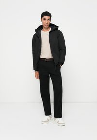 Zwarte gewatteerde jas met capuchon, beige trui, zwarte broek en witte sneakers met grijze accenten. Model staat tegen een effen achtergrond.