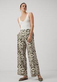 Bredde benmønstrede bukser i oliven og krem, med et intrikat design. Kombinert med en tettsittende hvit tank topp og slip-on sko.