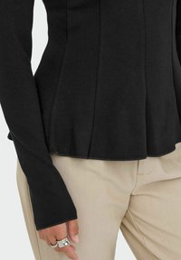 Black long-sleeve top with a peplum design, featuring a textured knit fabric. Paired with beige trousers and silver rings on a hand.