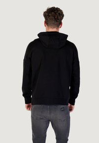 Felpa nera con cappuccio, dal fit rilassato e polsini a coste; mostrata da dietro, abbinata a jeans grigi in denim con dettagli minimi.